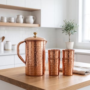 May include: A hammered copper pitcher with a matching lid and handle, accompanied by two stacked sets of hammered copper tumblers. The pitcher features a metallic finish and a cylindrical body, while the tumblers have a consistent hammered texture.