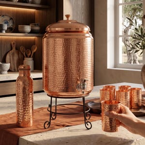 Puede incluir: Un gran dispensador de agua de cobre martillado con tapa y grifo de metal, apoyado sobre un soporte de metal negro. A su lado hay una botella de cobre martillado a juego y varios vasos de cobre martillado.