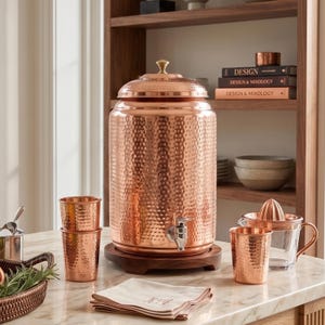 May include: A large hammered copper water dispenser with a matching lid and a silver-toned spigot, resting on a circular wooden base. The set includes two matching hammered copper tumblers and a glass citrus juicer with a copper handle.