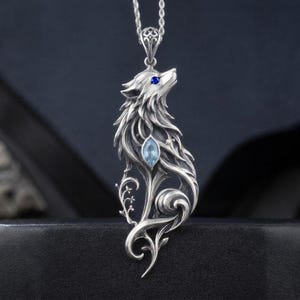 Puede incluir: Un colgante de plata con forma de cabeza de lobo estilizada, con un pequeño ojo de piedra preciosa azul y una piedra azul claro más grande de corte marquesa en el cuerpo. El diseño presenta intrincados detalles metálicos y motivos florales.
