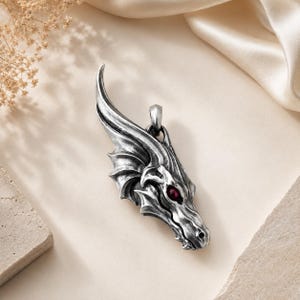 May include: A silver-toned metal pendant shaped like a dragon head with intricate scales and a single, prominent dark red gemstone set into the eye socket. The pendant features a curved horn or crest at the top and a small loop for a chain attachment.