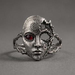 Puede incluir: Un anillo de metal plateado con un rostro esculpido de textura agrietada. Un ojo tiene una piedra preciosa roja oscura facetada, mientras que la otra cuenca está vacía. La banda está formada por estructuras metálicas orgánicas y ramificadas.