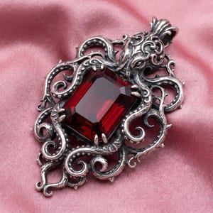 May include: A silver-toned pendant featuring an intricate octopus design with swirling tentacles surrounding a large, rectangular, deep red gemstone set in the center. The metalwork displays detailed textures and a dark, oxidized finish.