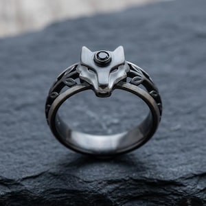 Könnte beinhalten: Ein silberfarbener Ring mit einem Wolfskopf-Design und einem schwarzen Edelsteinauge. Der Ring hat ein kompliziertes Blattmuster auf dem Band. Der Ring ist auf einer dunklen, strukturierten Oberfläche platziert.