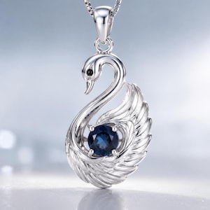 Può includere: Un ciondolo color argento a forma di cigno con piume dettagliate sulle ali e un piccolo occhio nero. Una gemma rotonda di colore blu intenso è incastonata al centro del corpo del cigno. Il ciondolo pende da una sottile catenina color argento.