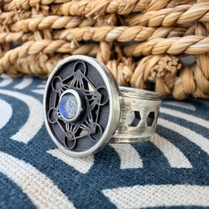 May include: A silver ring featuring a circular face with a black geometric Metatron's cube cutout design and a central round iridescent moonstone cabochon. The wide band has a brushed metal finish and a series of hexagonal cutouts along the side.