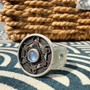 Op de afbeelding: Een zilverkleurige ring met een cirkelvormig voorvlak met een donker, verdiept geometrisch patroon rondom een kleine, ronde, iriserende blauwe cabochonsteen. De ringband heeft een geborstelde metalen afwerking en een zeshoekige uitsparing.