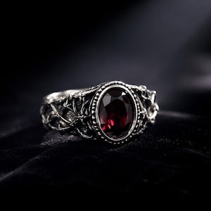 Puede incluir: Un anillo de plata con una piedra preciosa de color rojo intenso de corte ovalado engastada en un bisel con cuentas. La banda presenta un patrón de relieve texturizado similar a una enredadera que envuelve los lados con metal detallado.