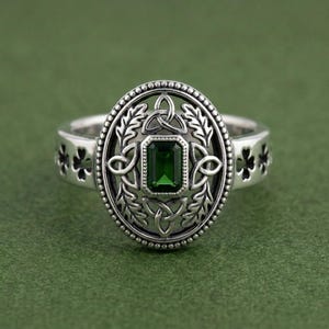 May include: A silver ring with an oval centerpiece featuring a green emerald-cut gemstone. The ring has Celtic knot and leaf designs, with clover cutouts on the band. The ring is set against a green background.