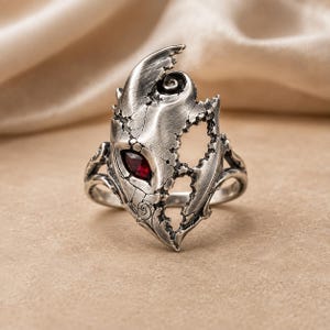 Può includere: Un anello color argento caratterizzato da un intricato design a forma di maschera usurata con una texture screpolata e una singola gemma rosso scuro taglio marquise incastonata al centro. La superficie metallica ha una finitura ossidata.