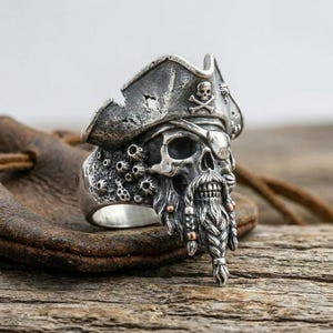 Op de afbeelding: Een zilveren ring met een gedetailleerde schedel die een piratenhoed met een doodskopembleem draagt. De schedel heeft een gevlochten baard met kleine metalen accenten en pokachtige texturen aan de zijkant van de ringband in geoxideerd zilver.
