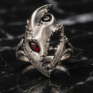 Puede incluir: Un anillo de tono plateado con una textura compleja, desgastada y agrietada, que presenta una piedra preciosa roja oscura de corte marquesa engastada en el centro. El metal muestra patrones en espiral y bordes irregulares.