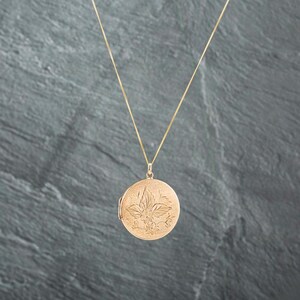 May include: A gold-toned circular locket pendant suspended from a delicate matching chain. The front surface of the locket features an intricate, etched floral design with detailed leaves and small blossoms, creating a textured, matte finish.