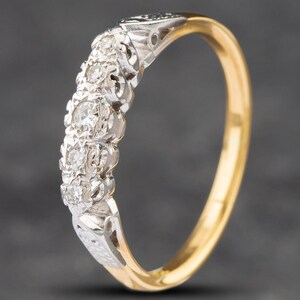 Peut inclure: Une bague en or jaune avec un sertissage argenté contenant cinq pierres rondes, claires et facettées disposées en ligne verticale. L'anneau est lisse et poli, tandis que le sertissage présente un travail du métal complexe et détaillé.