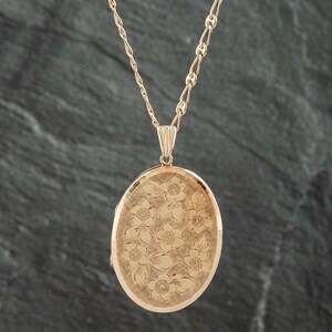 May include: An oval gold locket pendant suspended from a matching gold chain. The front surface of the locket features an intricate engraved pattern of multiple flowers and leaves with a textured background finish, creating a detailed floral design.