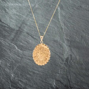 May include: A gold oval locket pendant suspended from a thin gold chain. The locket features an intricate engraved floral pattern on its front surface and is framed by a decorative scalloped edge, creating a textured and detailed appearance.