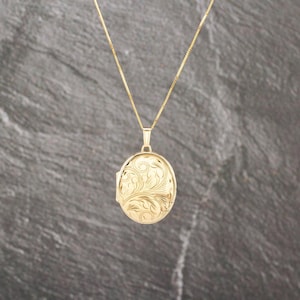 May include: A gold oval locket pendant suspended from a delicate gold chain. The locket features an intricate, engraved floral scrollwork pattern across its front surface, showcasing a polished metallic finish and a classic, symmetrical design.