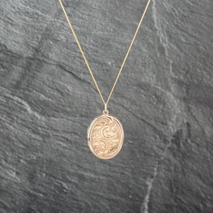 May include: A gold-toned oval locket pendant suspended from a delicate matching chain. The locket features an intricate, engraved swirling pattern across its front surface, creating a textured appearance that catches the light.