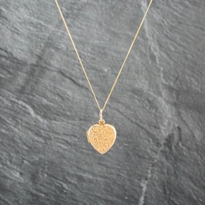 May include: A gold heart-shaped locket pendant suspended from a delicate gold chain. The surface of the heart locket features an intricate, embossed floral scrollwork pattern across the entire front face, creating a textured and detailed appearance.