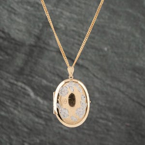 May include: An oval gold locket pendant suspended from a matching gold chain. The locket features a central dark oval inlay surrounded by an engraved silver-toned floral pattern on a gold-toned surface, creating a two-tone metallic appearance.