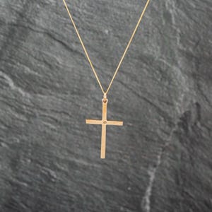 May include: A gold-toned cross pendant suspended from a thin, matching gold-toned chain. The cross features a simple, flat design with a small, singular circular accent at the center where the vertical and horizontal bars intersect.