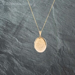 May include: A gold-toned oval locket pendant suspended from a delicate matching chain. The front surface of the locket features an intricate, symmetrical floral-inspired engraving centered on the polished metal face.