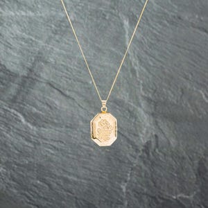 May include: A gold-toned locket pendant with an octagonal shape suspended from a thin gold-toned chain. The front surface of the locket features an engraved floral design with a central flower and leafy stems, showcasing a polished metallic finish.