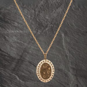 May include: A gold-toned oval locket pendant suspended from a matching twisted rope chain. The locket features an intricate filigree border surrounding a central oval panel engraved with detailed swirling floral patterns in a metallic finish.