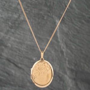 May include: A gold-toned oval locket pendant suspended from a matching delicate gold-toned chain. The front surface of the locket features an intricate engraved floral pattern on one side, while the remaining surface is smooth and polished.