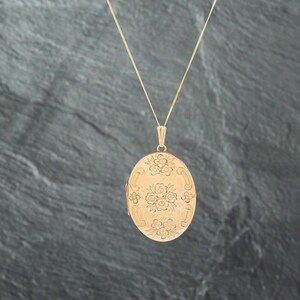 May include: An oval gold locket pendant suspended from a thin gold chain. The front surface of the locket features an engraved floral pattern with clusters of small flowers arranged in the center and at the top and bottom of the oval shape.