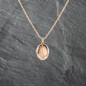 May include: A rose gold-toned oval locket pendant suspended from a matching twisted rope chain. The locket features an engraved decorative leaf-like pattern on the left side, while the remaining surface is smooth and polished with a metallic finish.
