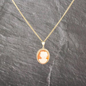 Könnte beinhalten: Eine goldene Halskette mit einem ovalen Anhänger, der ein geschnitztes Kamee-Profil einer Person zeigt. Der Anhänger ist von einem strukturierten Goldrand umgeben und hängt an einer feinen Goldgliederkette in hellorangen und weißen Farben.