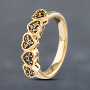 May include: A gold ring featuring a series of five connected heart-shaped settings along the top. Each heart-shaped setting contains three small, round, black stones. The band is smooth and polished, showcasing a continuous yellow gold metal finish.