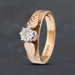 May include: A rose gold ring featuring a textured bark-like finish on the band, accented by two engraved leaf motifs and a central silver-toned flower setting containing a single small round clear gemstone at its center point.