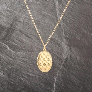 May include: A gold-toned oval locket pendant suspended from a matching delicate link chain. The front surface of the locket features a raised, repeating diamond-shaped grid pattern with small dots at each intersection, creating a textured appearance.