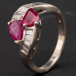May include: A silver-toned metal ring featuring two pear-cut deep red rubies set side-by-side. The band is accented with a row of clear, rectangular baguette-cut stones along the shank, creating a bypass design with a polished metallic finish.