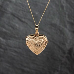 May include: A gold-toned heart-shaped locket pendant suspended from a delicate matching chain. The locket features an intricate engraved pattern on its front surface, consisting of fine diagonal lines and a stylized leaf motif in a polished finish.