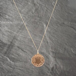 May include: A gold-toned circular locket pendant suspended from a thin matching chain. The locket features an intricate embossed floral scrollwork pattern surrounding a smooth, shield-shaped center panel on its front surface.