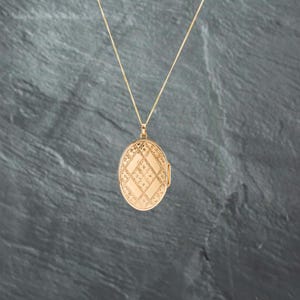 May include: An oval gold locket pendant suspended from a thin gold chain. The locket features a textured surface with a repeating diamond pattern, where alternating sections display a smooth finish and a detailed, intricate floral-like engraving.