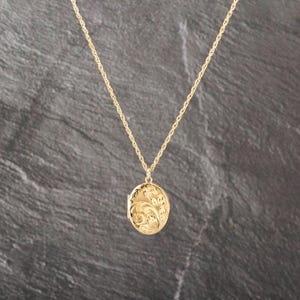 May include: A gold-toned oval locket pendant suspended from a delicate matching gold-toned rope chain. The locket features an intricate, embossed floral scrollwork pattern across its entire front surface, creating a textured and detailed appearance.