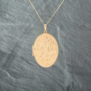 May include: An oval gold locket pendant suspended from a thin gold chain. The front surface of the locket features an intricate, embossed floral scrollwork pattern covering the entire face, with a visible hinge on the left side of the oval frame.