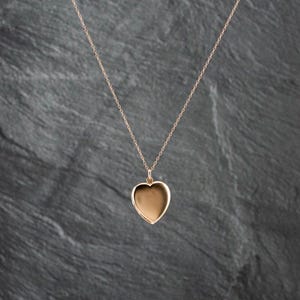 May include: A rose gold heart-shaped locket pendant suspended from a delicate matching rose gold chain. The locket features a smooth, polished surface with a simple, classic design, reflecting light across its rounded, symmetrical heart shape.