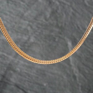Puede incluir: Un collar de cadena de espiga en tono oro rosa con un patrón de eslabones planos, flexibles y estrechamente tejidos que crea una superficie lisa y continua. El metal tiene un acabado pulido que refleja la luz sobre los eslabones.