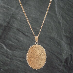 May include: A gold-toned oval locket pendant suspended from a matching delicate link chain. The locket features an intricate, engraved swirling floral pattern across its entire front surface, with a scalloped edge detail surrounding the oval shape.