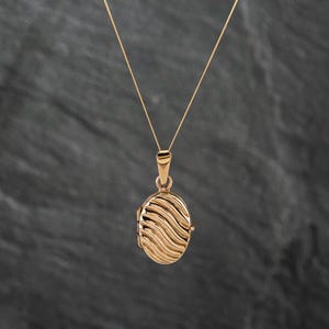 May include: A rose gold oval locket pendant suspended from a delicate matching chain. The locket features a textured surface with a series of raised, diagonal wavy lines that create a rhythmic pattern across the entire front face of the piece.