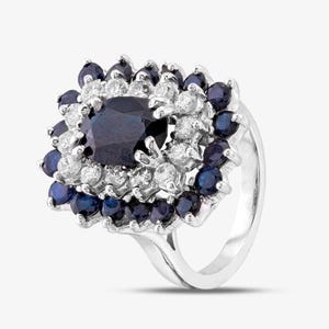Peut inclure: Une bague argentée ornée d'un grand saphir bleu foncé de taille ovale au centre, entouré d'un halo de petits diamants blancs ronds et d'une bordure extérieure de saphirs bleus ronds disposés en grappe sur un anneau en métal poli.