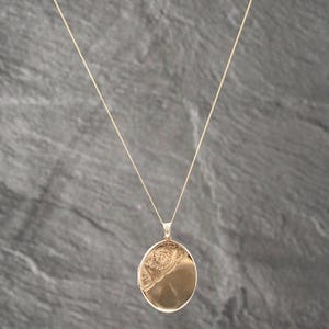May include: A gold-toned oval locket pendant suspended from a thin matching chain. The locket features a smooth, polished surface on the lower right side and an intricate, embossed scrollwork pattern covering the upper left portion of the face.