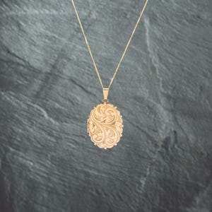 May include: A gold-toned oval locket pendant suspended from a delicate matching chain. The locket features an intricate, embossed floral scrollwork pattern across its entire front surface, creating a textured and detailed appearance.
