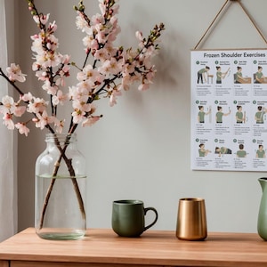 K&ouml;nnte beinhalten: Eine klare Glasvase mit rosa Kirschbl&uuml;tenzweigen, eine matte olivgr&uuml;ne Tasse, ein metallisch goldener Becher und ein matter olivgr&uuml;ner Krug. Ein wei&szlig;es Wandplakat mit dem Titel Frozen Shoulder Exercises zeigt zw&ouml;lf illustrierte physiotherapeutische &Uuml;bungen.
