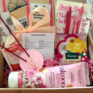 Könnte beinhalten: Eine Geschenkbox mit einem in beigem Papier verpackten Buch mit pfirsichfarbenem Band, einer Buchbewertungskarte, einem rosa Räucherstäbchenhalter mit zwei Stäbchen, einem Yogi-Teebeutel, Jean & Len Mandel-Duschbalsam und einer Sonnenblumen-Grußkarte.